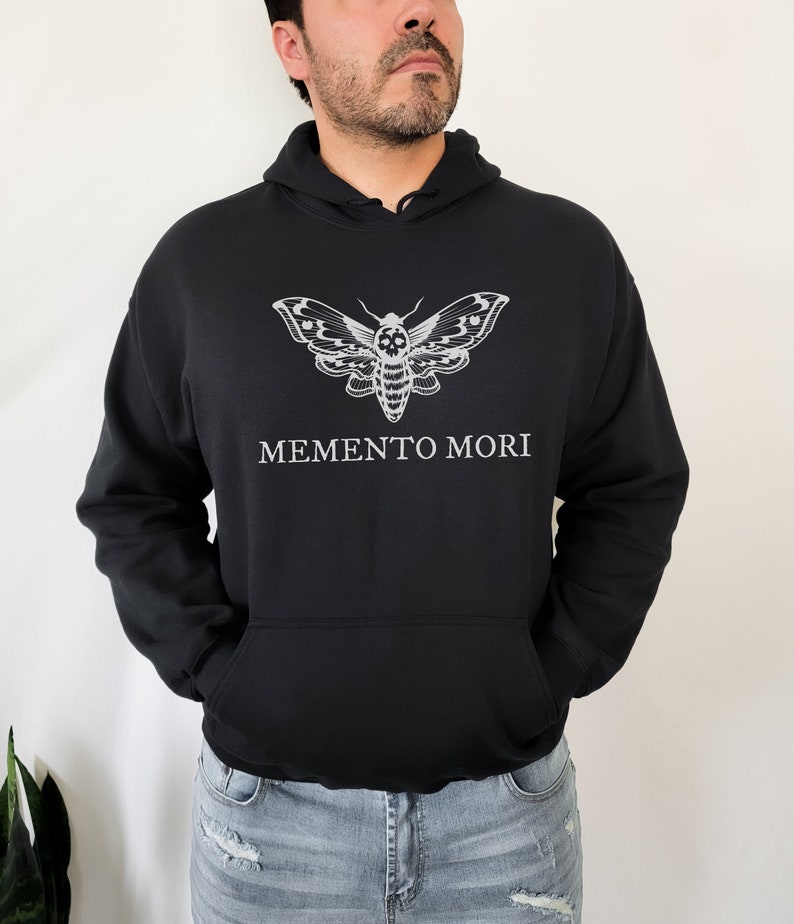 K&ouml;nnte beinhalten: Schwarzer Kapuzenpullover mit einem wei&szlig;en Totenkopf-Motten-Motiv und dem Text "MEMENTO MORI" auf der Vorderseite.