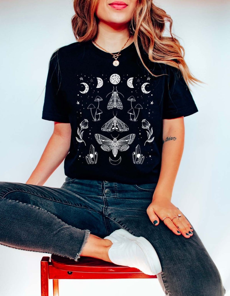 Puede incluir: Una camiseta negra con un dise&ntilde;o gr&aacute;fico blanco que presenta fases lunares, polillas, hongos y cristales.