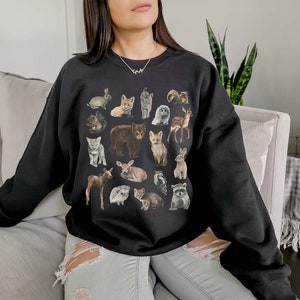 May include: A black sweatshirt featuring a graphic print of various woodland animals, including a bear, fox, deer, rabbit, and raccoon. The animals are depicted in a cartoon style and are arranged in a grid pattern.