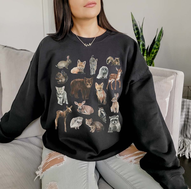 May include: A black sweatshirt featuring a graphic print of various woodland animals, including a bear, fox, deer, rabbit, and raccoon. The animals are depicted in a cartoon style and are arranged in a grid pattern.