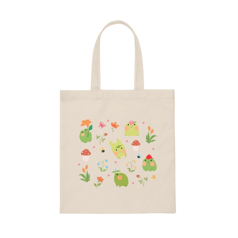 May include: A beige tote bag with a design of green frogs, flowers, mushrooms, and bees. The frogs are wearing flowers and hats.