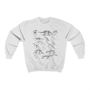 May include: White sweatshirt with a black and white illustration of dinosaur skeletons. The skeletons are arranged in rows and include a variety of dinosaurs, such as Stegosaurus, Triceratops, and Brachiosaurus.