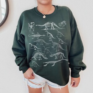 May include: A dark green sweatshirt with a white print of dinosaur skeletons. The print features various dinosaurs, including a Tyrannosaurus Rex, Triceratops, and Stegosaurus.