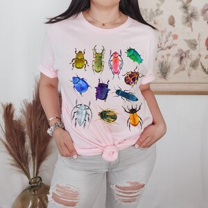 May include: A pink t-shirt with a colorful beetle design. The beetles are in various shades of green, blue, red, yellow, and purple.