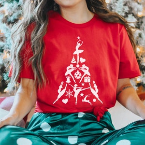 May include: A red t-shirt with a white design of a Christmas tree made up of ballerinas. The tree is decorated with ornaments, snowflakes, stars, and a reindeer.