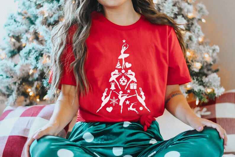 May include: A red t-shirt with a white design of a Christmas tree made up of ballerinas. The tree is decorated with ornaments, snowflakes, stars, and a reindeer.