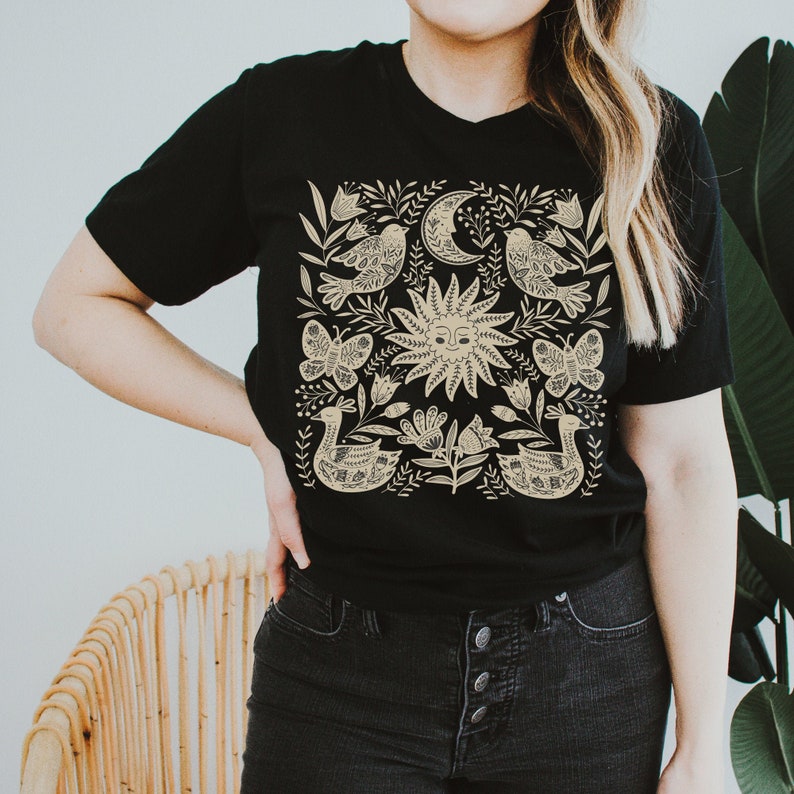 May include: A black t-shirt with a cream-colored design featuring a sun, moon, birds, butterflies, and flowers. The design is centered on the shirt and is surrounded by a black border.