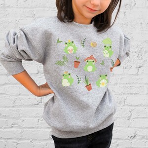 K&ouml;nnte beinhalten: Ein graues Sweatshirt mit einem niedlichen Frosch-Design. Die Fr&ouml;sche tragen H&uuml;te und halten Blumen. Das Sweatshirt hat einen Rundhalsausschnitt und lange &Auml;rmel.
