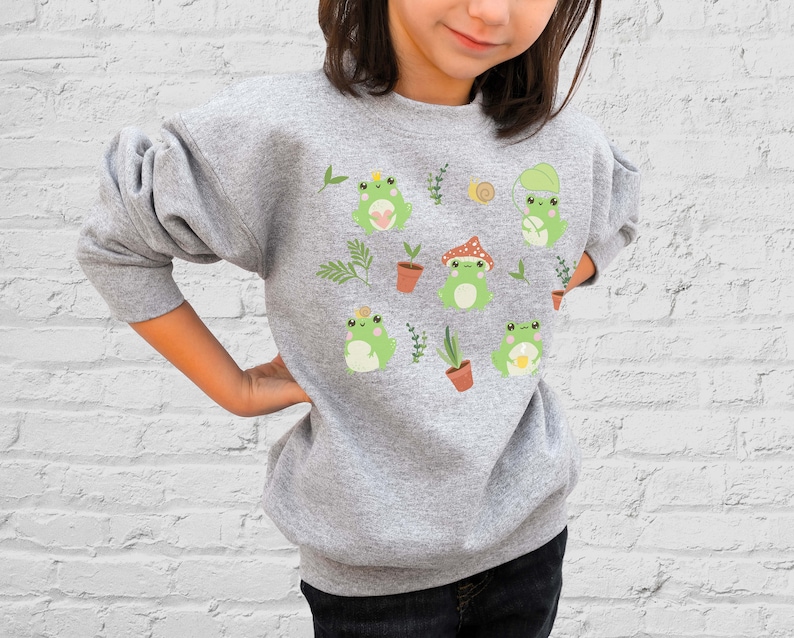 K&ouml;nnte beinhalten: Ein graues Sweatshirt mit einem niedlichen Frosch-Design. Die Fr&ouml;sche tragen H&uuml;te und halten Blumen. Das Sweatshirt hat einen Rundhalsausschnitt und lange &Auml;rmel.