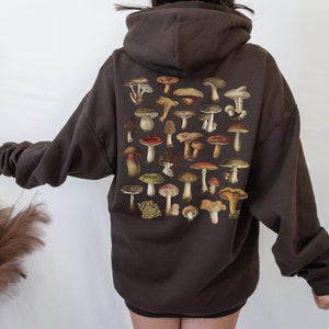 May include: A dark brown hoodie featuring a collection of various mushroom illustrations on the back. The mushrooms are depicted in a range of colors, including brown, red, and green. The hoodie has a relaxed fit and a hood.
