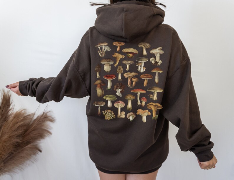 May include: A dark brown hoodie featuring a collection of various mushroom illustrations on the back. The mushrooms are depicted in a range of colors, including brown, red, and green. The hoodie has a relaxed fit and a hood.