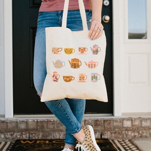 May include: A white canvas tote bag with a floral pattern of teapots and teacups. The bag has two handles and is being held by a person.