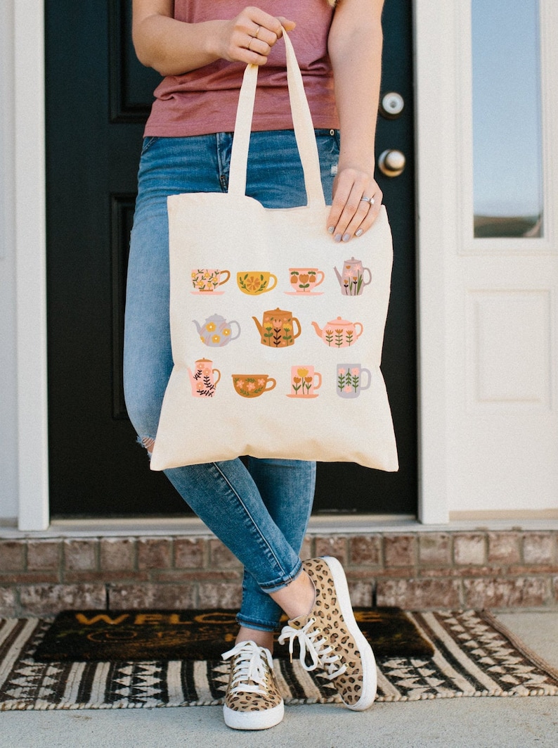 May include: A white canvas tote bag with a floral pattern of teapots and teacups. The bag has two handles and is being held by a person.