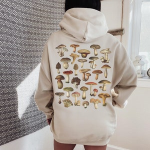 May include: Beige hoodie featuring a collection of various mushroom illustrations in shades of brown, red, and green. The mushrooms are arranged on the back of the hoodie. The hoodie has a drawstring hood and long sleeves.