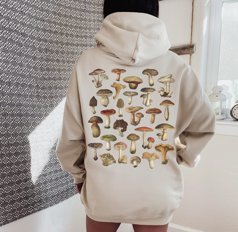 May include: Beige hoodie featuring a collection of various mushroom illustrations in shades of brown, red, and green. The mushrooms are arranged on the back of the hoodie. The hoodie has a drawstring hood and long sleeves.