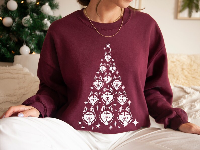 Puede incluir: Una sudadera color burdeos con un dise&ntilde;o de &aacute;rbol de Navidad blanco con m&uacute;ltiples caras de perros husky. El &aacute;rbol est&aacute; decorado con estrellas blancas.