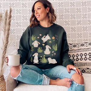 May include: A dark green sweatshirt with a white bunny rabbit print. The bunnies are surrounded by green grass and small white flowers.