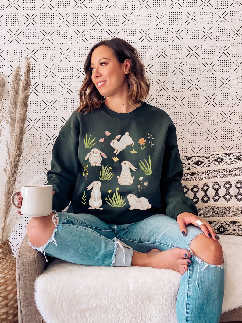 May include: A dark green sweatshirt with a white bunny rabbit print. The bunnies are surrounded by green grass and small white flowers.