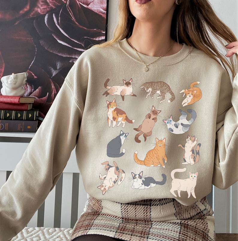 May include: A beige sweatshirt with a graphic print of various cats in different poses. The cats are all in shades of brown, white, and gray.