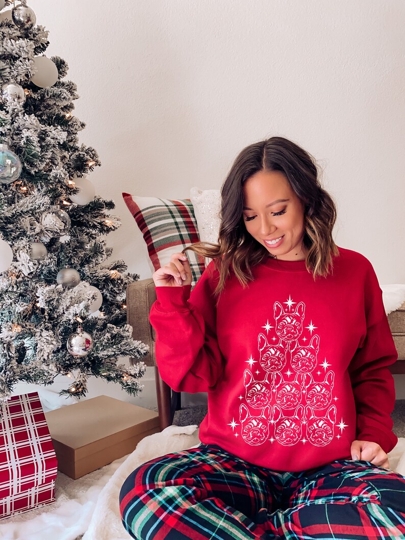 May include: A red Christmas sweater with a white design of a Christmas tree made of French bulldog faces. The sweater is worn by a person sitting on a floor with a plaid pattern.