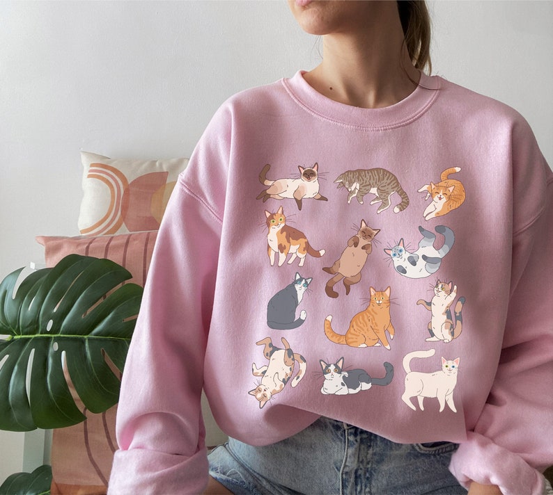 May include: A pink sweatshirt with a graphic print of various cats in different poses. The cats are all in shades of brown, white, gray, and black.