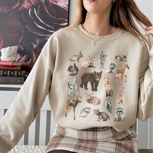May include: A beige sweatshirt featuring a graphic print of assorted woodland animals, such as a bear, deer, fox, rabbit, raccoon, and owl. The animals are illustrated in a cartoon style and are positioned in a circular arrangement.