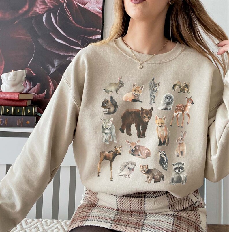 May include: A beige sweatshirt featuring a graphic print of assorted woodland animals, such as a bear, deer, fox, rabbit, raccoon, and owl. The animals are illustrated in a cartoon style and are positioned in a circular arrangement.