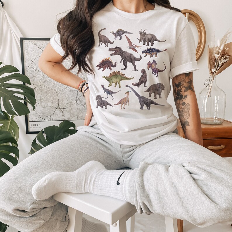 May include: A white t-shirt with a colorful dinosaur print. The shirt features a variety of dinosaurs, including a Tyrannosaurus Rex, a Triceratops, and a Stegosaurus.