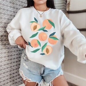 K&ouml;nnte beinhalten: Wei&szlig;er Pullover mit einem Pfirsich-Design. Die Pfirsiche sind mit gr&uuml;nen Bl&auml;ttern illustriert und haben einen pfirsichfarbenen Hintergrund.