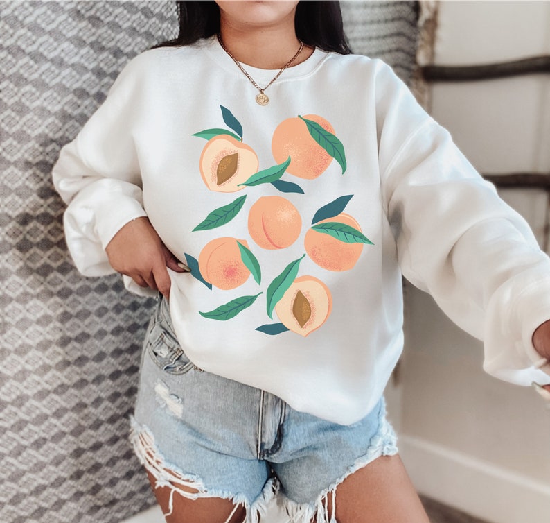 K&ouml;nnte beinhalten: Wei&szlig;er Pullover mit einem Pfirsich-Design. Die Pfirsiche sind mit gr&uuml;nen Bl&auml;ttern illustriert und haben einen pfirsichfarbenen Hintergrund.