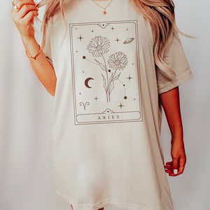 May include: A beige oversized t-shirt with a graphic print of a floral design with a crescent moon, stars, and the zodiac symbol for Aries. The text "ARIES" is printed below the design.