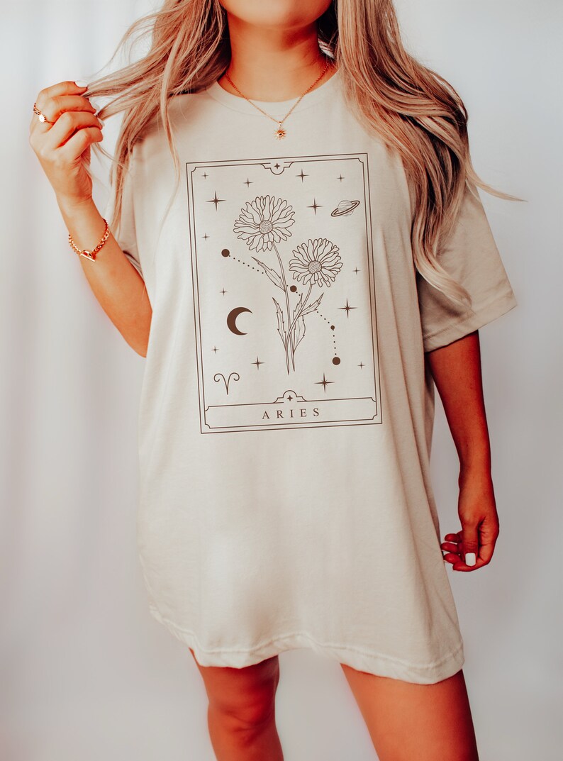 May include: A beige oversized t-shirt with a graphic print of a floral design with a crescent moon, stars, and the zodiac symbol for Aries. The text "ARIES" is printed below the design.