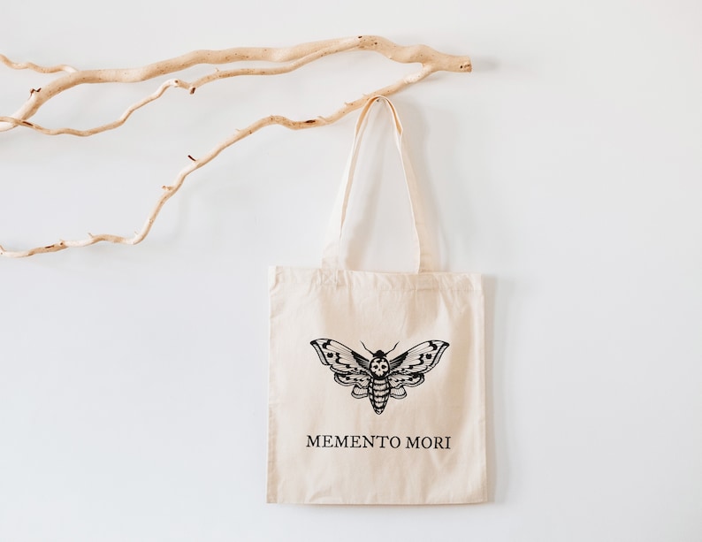May include: A beige tote bag with black straps and a black moth design. The bag has the text "MEMENTO MORI" printed on the front.