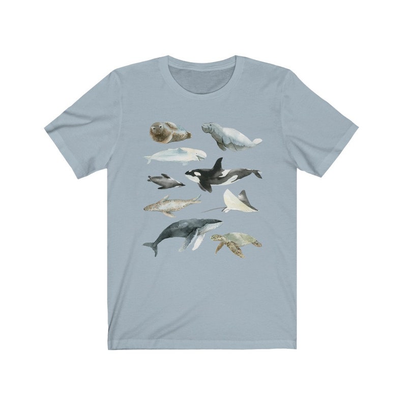 K&ouml;nnte beinhalten: Ein hellblaues T-Shirt mit einer Aquarell-Illustration verschiedener Meerestiere, darunter ein Wal, ein Delfin, eine Meeresschildkr&ouml;te, ein Seekuh, eine Robbe und ein Rochen.