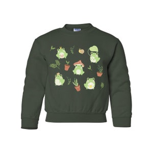 K&ouml;nnte beinhalten: Ein dunkelgr&uuml;ner Pullover mit einem Muster aus gr&uuml;nen Fr&ouml;schen, Pflanzen und Pilzen. Die Fr&ouml;sche tragen Kronen und halten Blumen.