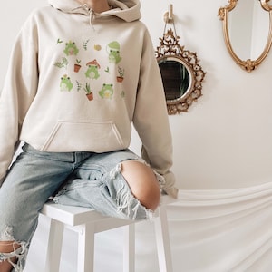 May include: A beige hoodie with a graphic print of frogs, plants, and mushrooms. The hoodie has a drawstring hood and a kangaroo pocket.