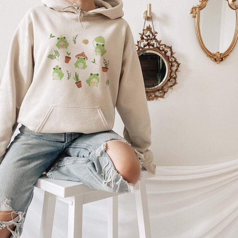 May include: A beige hoodie with a graphic print of frogs, plants, and mushrooms. The hoodie has a drawstring hood and a kangaroo pocket.