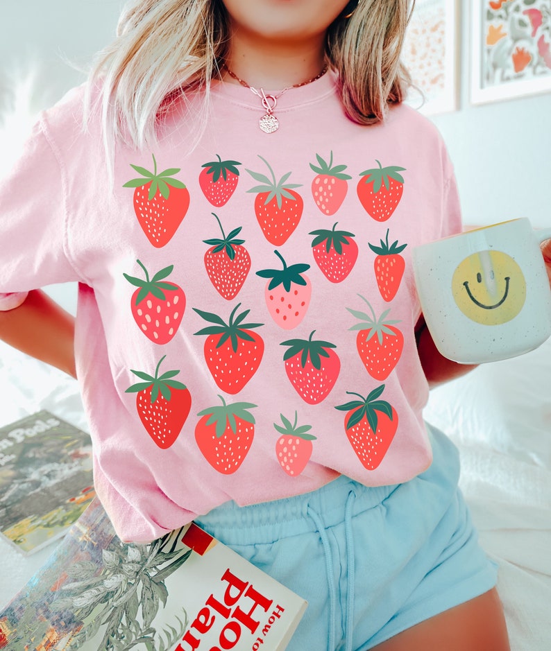 Puede incluir: Una camiseta rosa con un estampado gr&aacute;fico de fresas rojas. La camiseta tiene cuello redondo y mangas cortas.