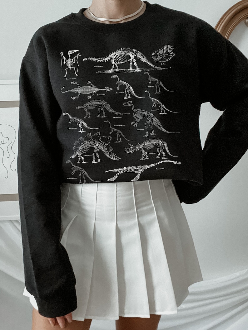 May include: Black cropped sweatshirt with a white print of dinosaur skeletons. The sweatshirt is paired with a white pleated skirt.