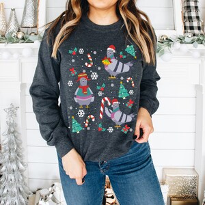 May include: A grey sweatshirt with a Christmas-themed design featuring pigeons wearing Santa hats and holding gifts. The design also includes candy canes, snowflakes, and Christmas trees.