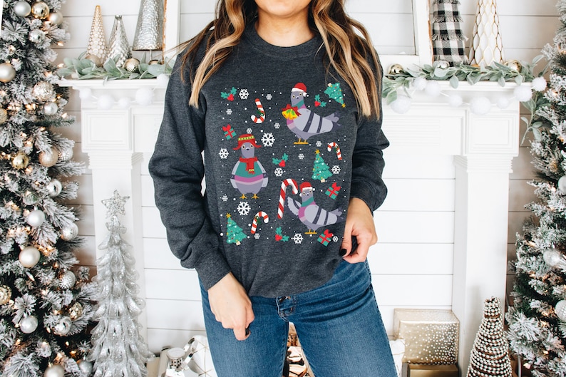 May include: A grey sweatshirt with a Christmas-themed design featuring pigeons wearing Santa hats and holding gifts. The design also includes candy canes, snowflakes, and Christmas trees.