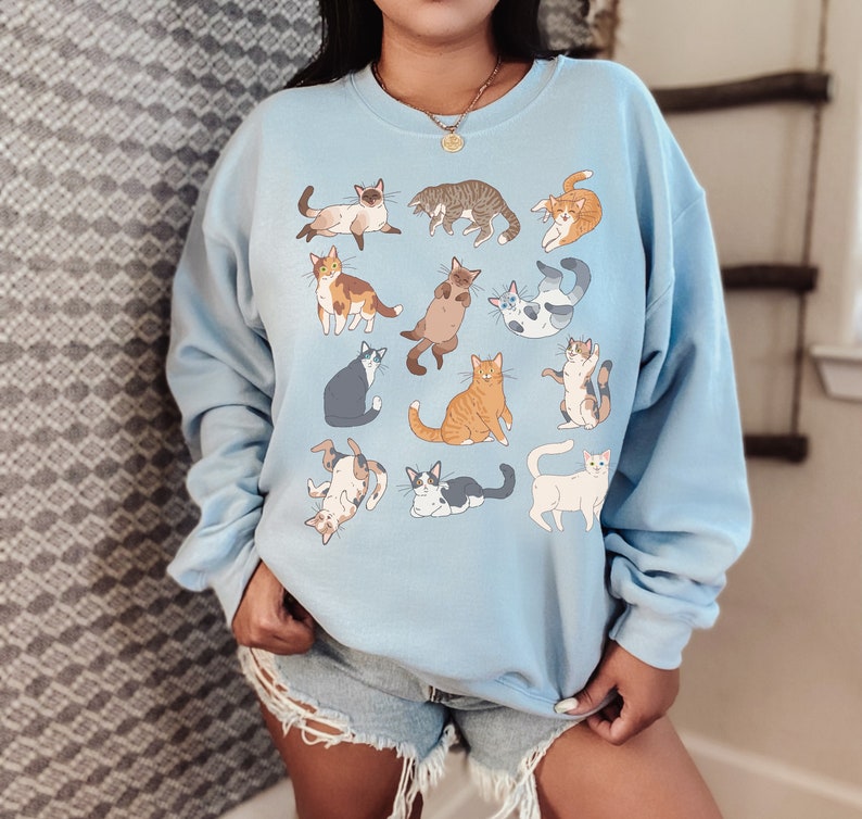 May include: A light blue sweatshirt with a graphic print of 12 cats in various poses. The cats are all different colors and have different patterns.