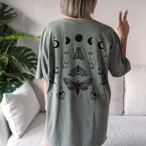 Op de afbeelding: Een saliegroen T-shirt met een zwarte grafische print van motten, paddenstoelen, kristallen en manen.