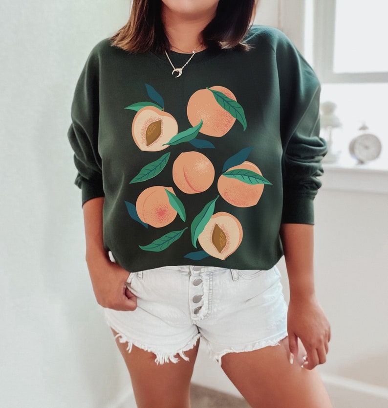 K&ouml;nnte beinhalten: Ein dunkelgr&uuml;ner Pullover mit einem Grafikdruck von Pfirsichen und Bl&auml;ttern. Der Pullover hat einen Rundhalsausschnitt und lange &Auml;rmel.