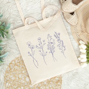 May include: A white canvas tote bag with blue floral line drawings. The bag has two handles and is lying on a white fuzzy surface.