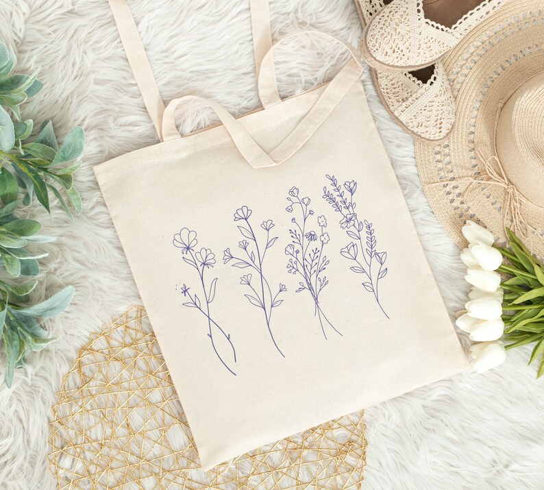 May include: A white canvas tote bag with blue floral line drawings. The bag has two handles and is lying on a white fuzzy surface.