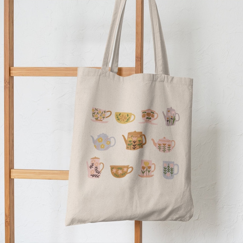 May include: A white canvas tote bag with a floral pattern of teapots and teacups in shades of blue, yellow, pink, and green.