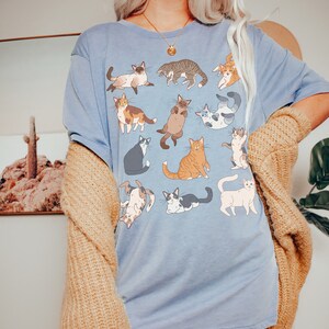 May include: A light blue t-shirt featuring a collection of cartoon cats in various poses. The cats are in different colors and patterns, including tabby, calico, and Siamese. The shirt is worn with a tan cardigan.
