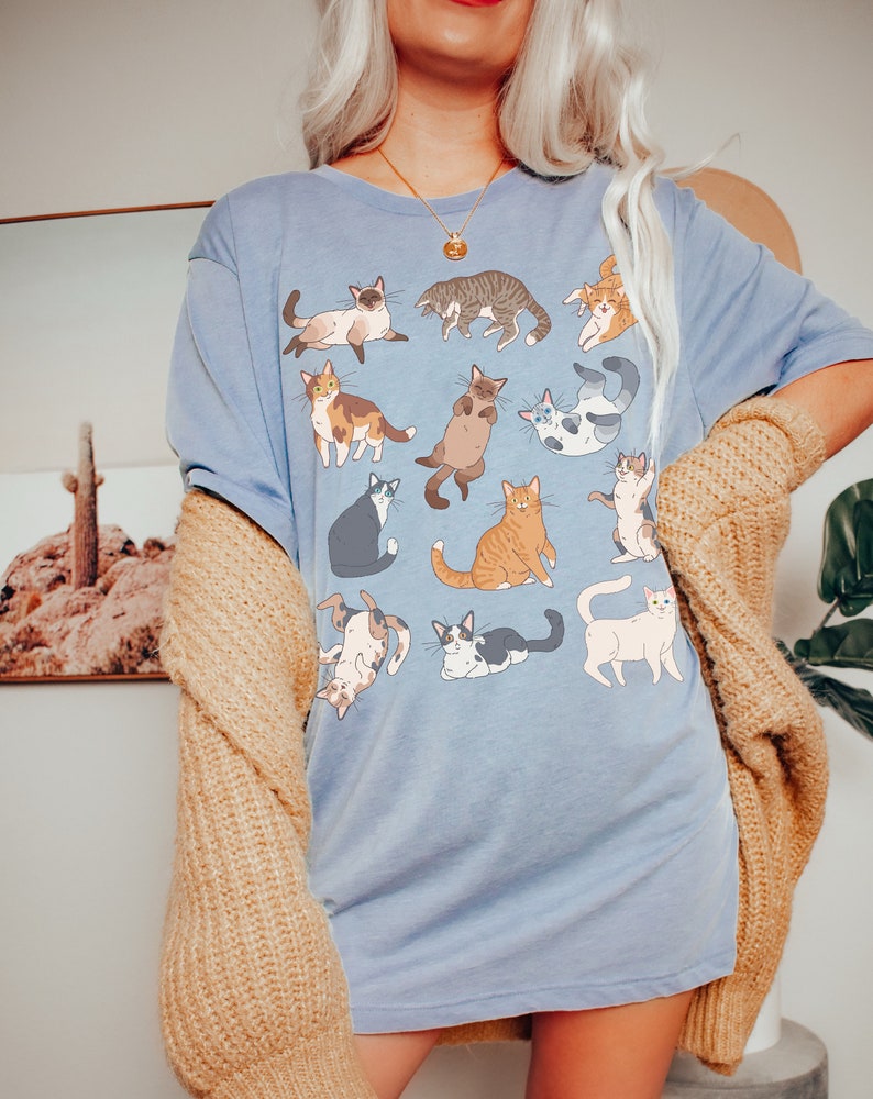 May include: A light blue t-shirt featuring a collection of cartoon cats in various poses. The cats are in different colors and patterns, including tabby, calico, and Siamese. The shirt is worn with a tan cardigan.
