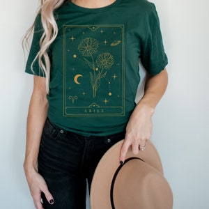 May include: A green t-shirt with a gold graphic of a flower with a crescent moon and stars. The graphic is in a rectangular frame with the text "ARIES" at the bottom.